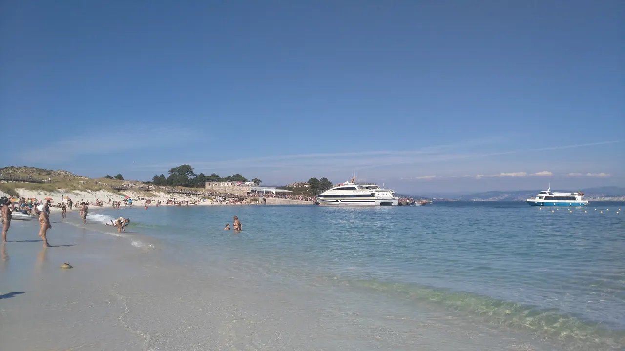 Barcos de pasajeros atracados en el muelle de las Islas Cíes, vistos desde la playa del embarcadero, con bañistas en la orilla