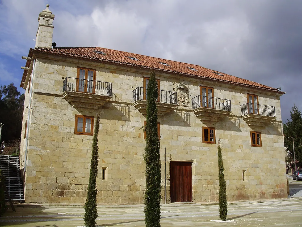 Pazo de Mos al sol, con su fachada de granito, balcones de hierro y tres cipreses delante, en A Rúa (Mos)