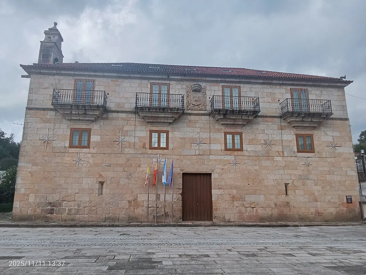 Fachada principal del Pazo de Mos en un día nublado, con cuatro balcones de hierro, el escudo en el centro y banderas junto a la puerta