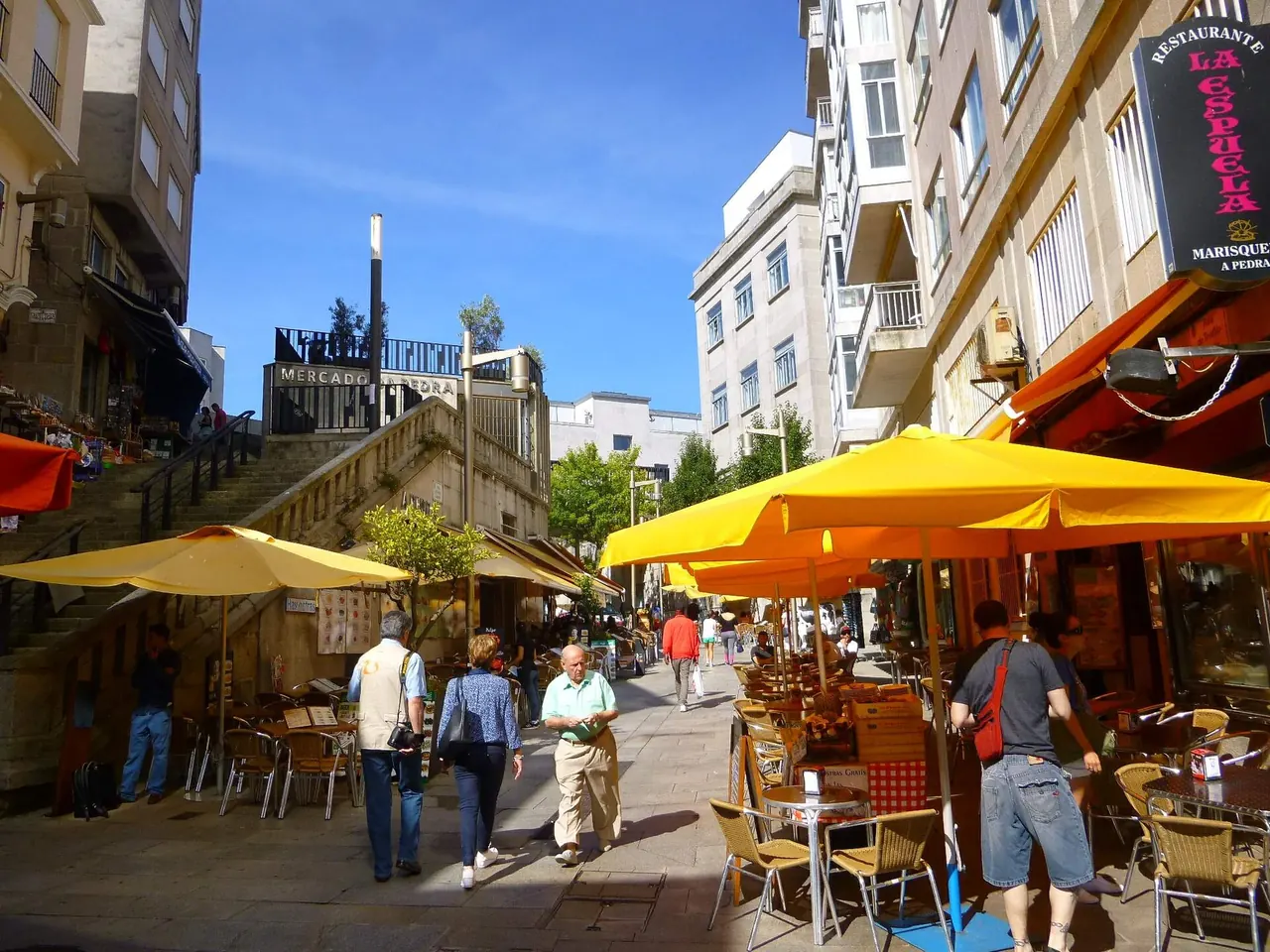 Calle peatonal junto al mercado de A Pedra, en Vigo, con sombrillas amarillas, terrazas y gente paseando