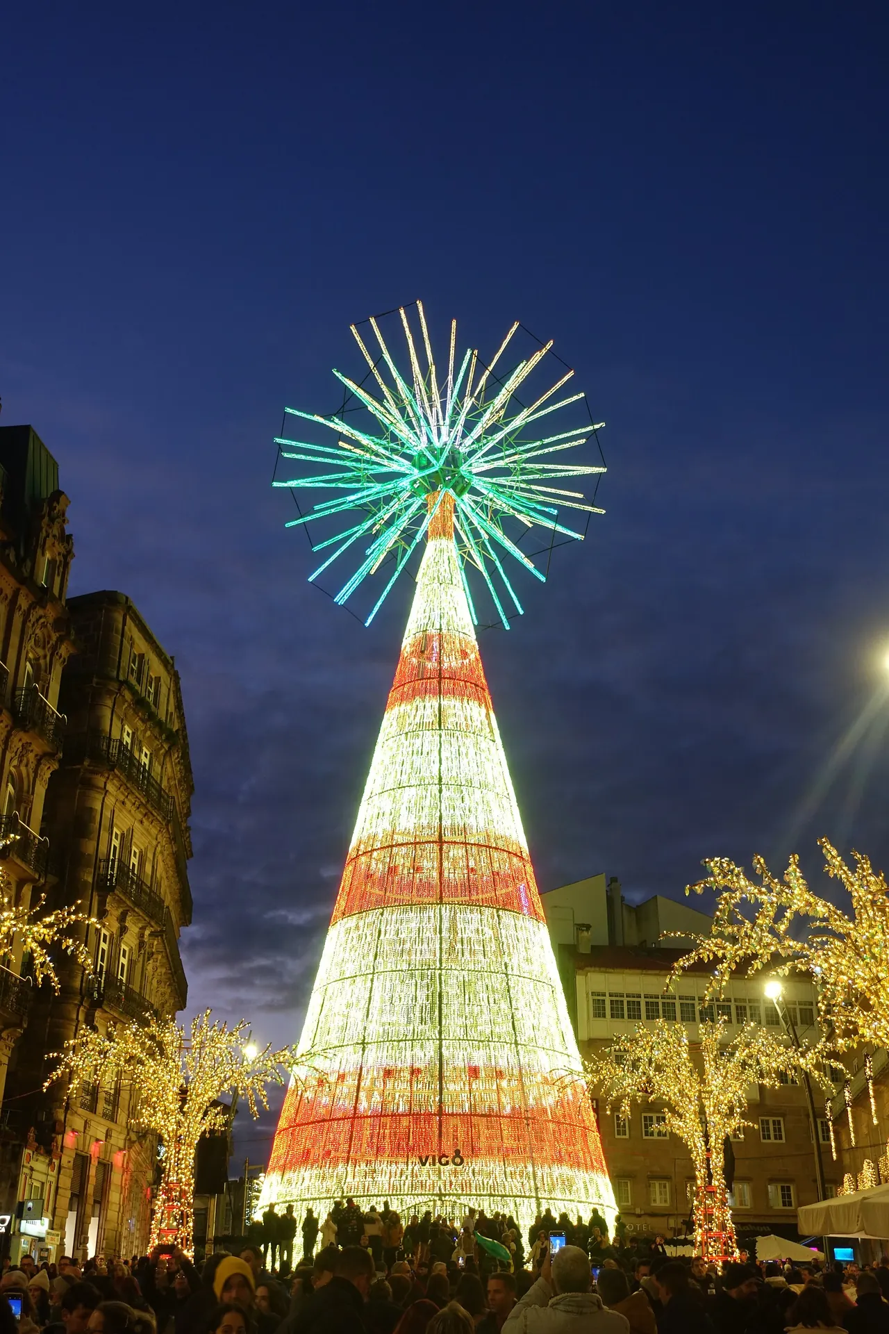 Árbol de Navidad gigante de luces blancas y rojas en Vigo, con una estrella de rayos verdes en lo alto, rodeado de gente de noche en la Navidad de 2024
