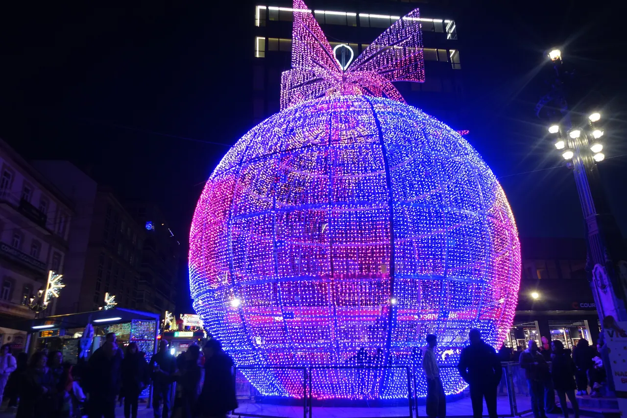 Bola de Navidad gigante de luces azules y rosas, con un lazo en lo alto, y gente alrededor de noche en Vigo en la Navidad de 2024