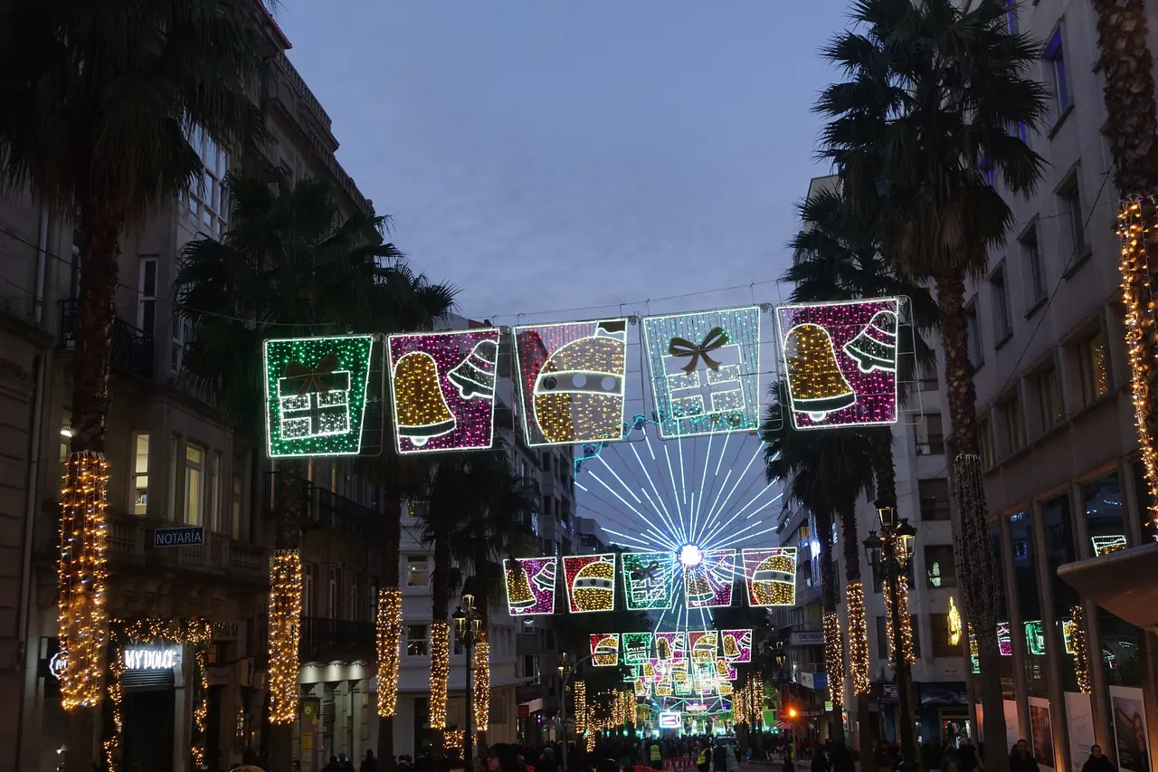 Calle de Vigo de noche en la Navidad de 2024, con arcos de luces de regalos y campanas, una rueda de luces al fondo y palmeras envueltas en luces