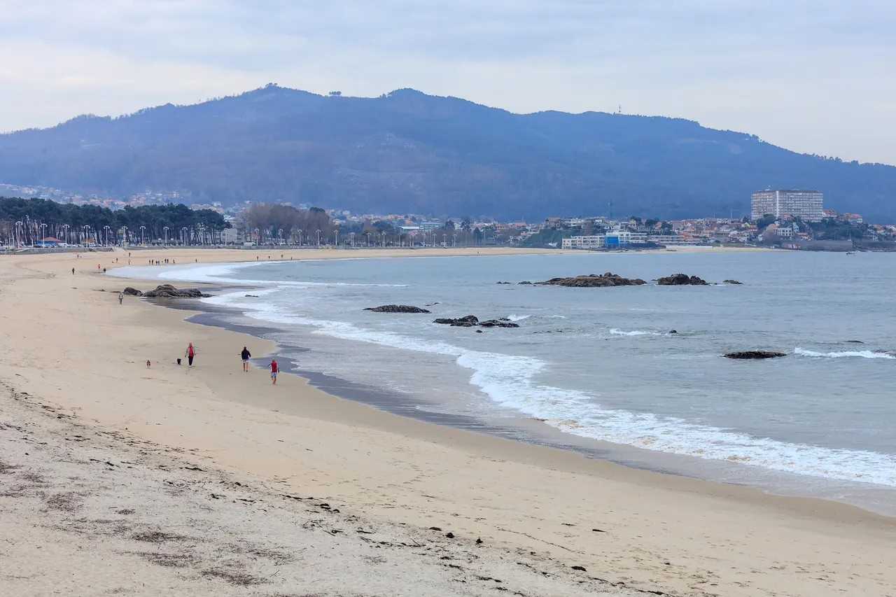 Playa de Samil, en Vigo, un día nublado: arena ancha, rocas en la orilla, algunos paseantes y el monte al fondo