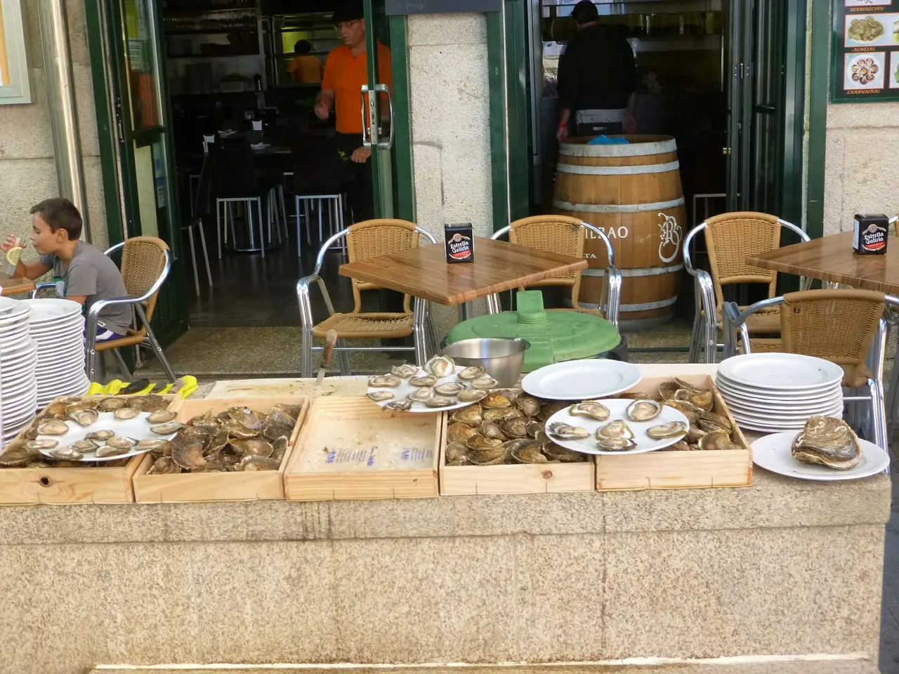 Bandejas de ostras abiertas sobre cajas de madera en un puesto de la calle de las ostras de Vigo, con mesas de terraza detrás